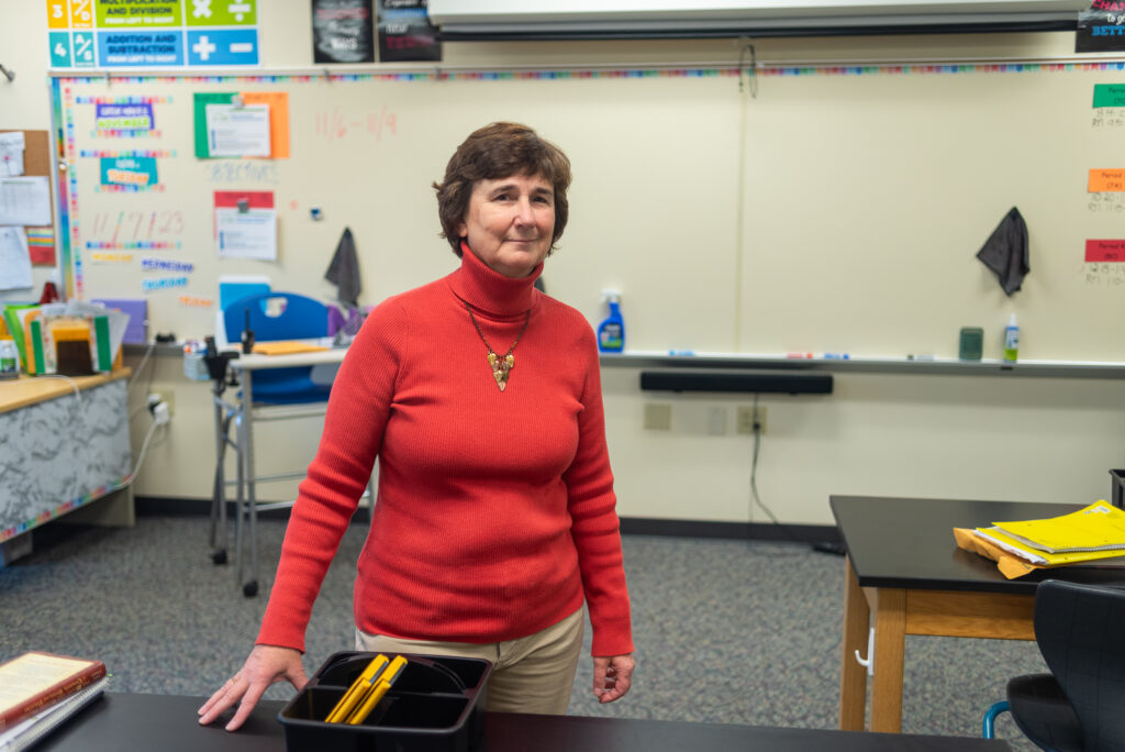 Kim stands in an empty classroom, looking into the camera. Her hand rests on a desk. 