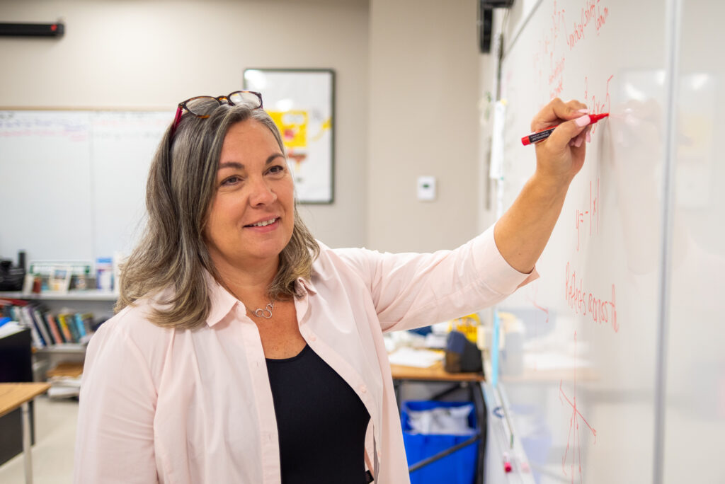 Jenn is standing at the whiteboard in her classroom, writing on the board with a red marker.