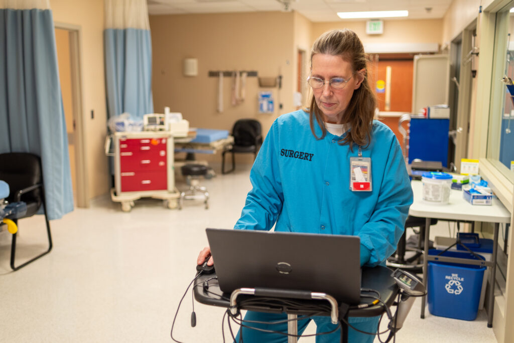 Shirley stands in an exam room, working at a laptop on a rolling station. She is wearing surgical scrubs.