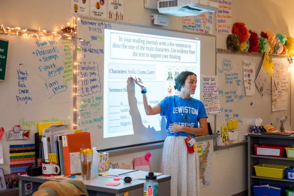 Kennedy stands at the white board, teaching a lesson. She is pointing to text projected on the white board directing the students to describe one of the main characters in the book they are reading.